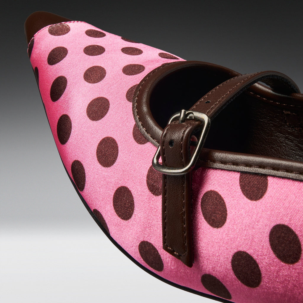 A close-up of a pink pointed-toe shoe with dark brown polka dots, a dark brown strap, and a silver buckle.