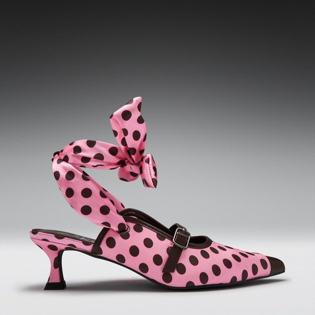 A pink pointed-toe heeled mule with black polka dots, featuring a matching polka-dot ankle strap tied in a large bow, photographed on a dark gray gradient studio background.