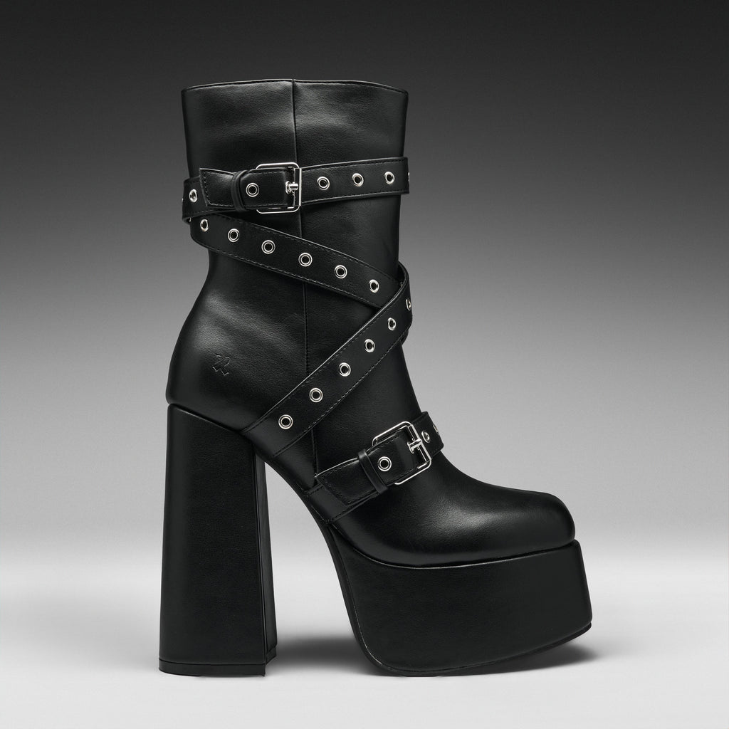 A black leather ankle boot with a chunky block heel, platform sole, and multiple decorative straps adorned with silver studs and buckles, shown against a dark gray gradient background.