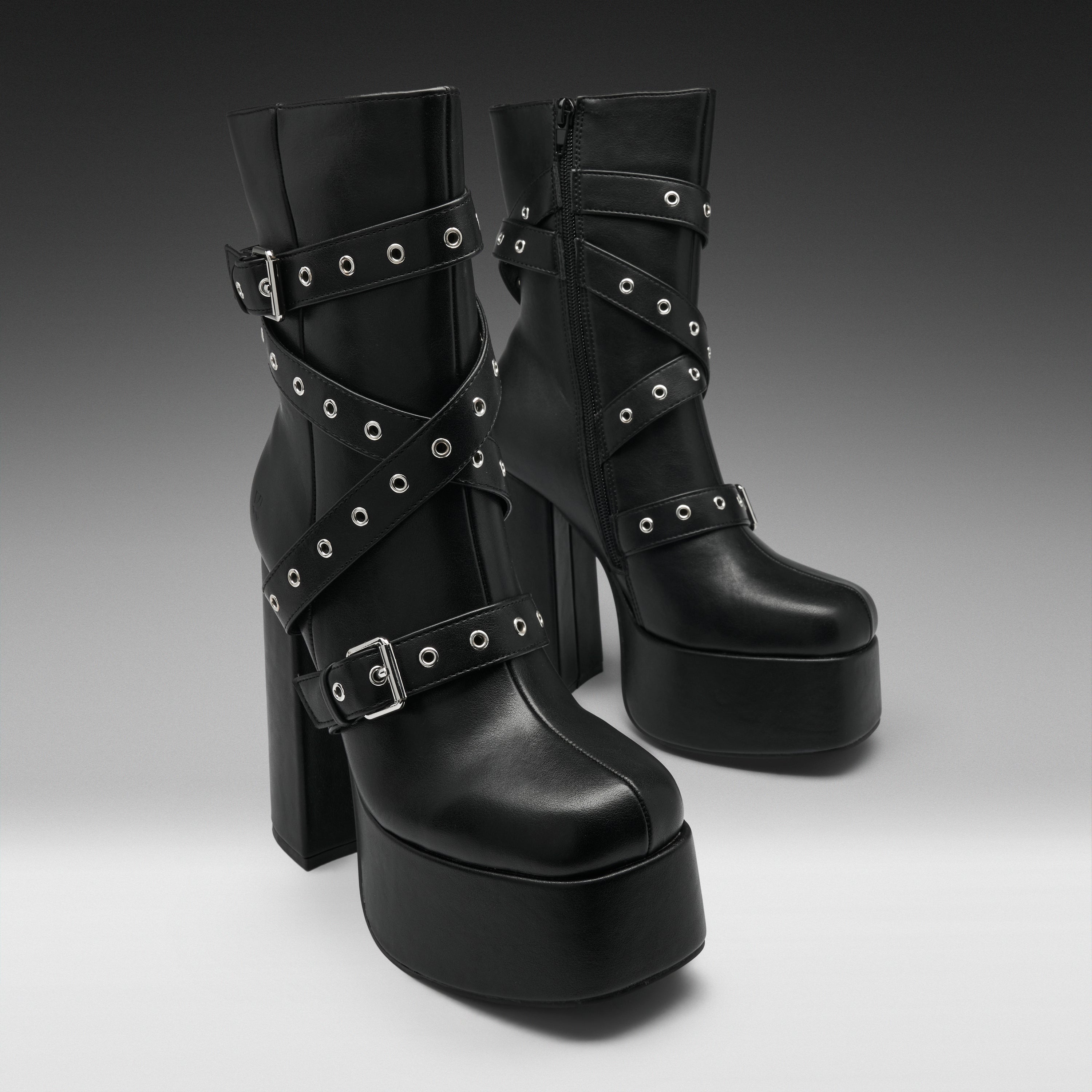 A pair of black platform ankle boots with high block heels, decorated with multiple crisscrossing straps and silver-tone metal grommets, shown against a dark-to-light gray gradient studio background.