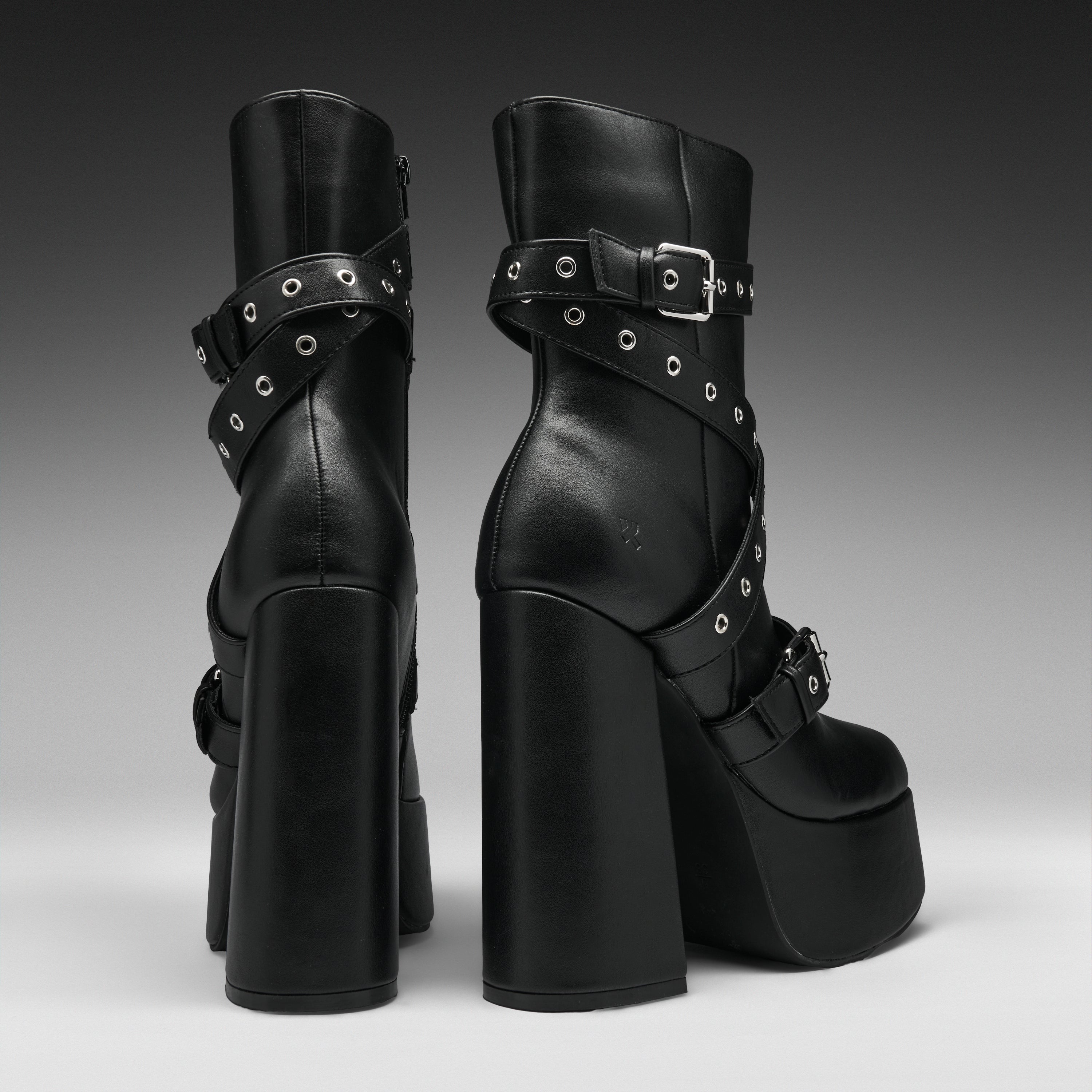 A pair of black platform ankle boots with chunky block heels, double platform soles, and multiple decorative straps with silver-tone grommets and buckles, shown from the back and side against a dark gray gradient background.