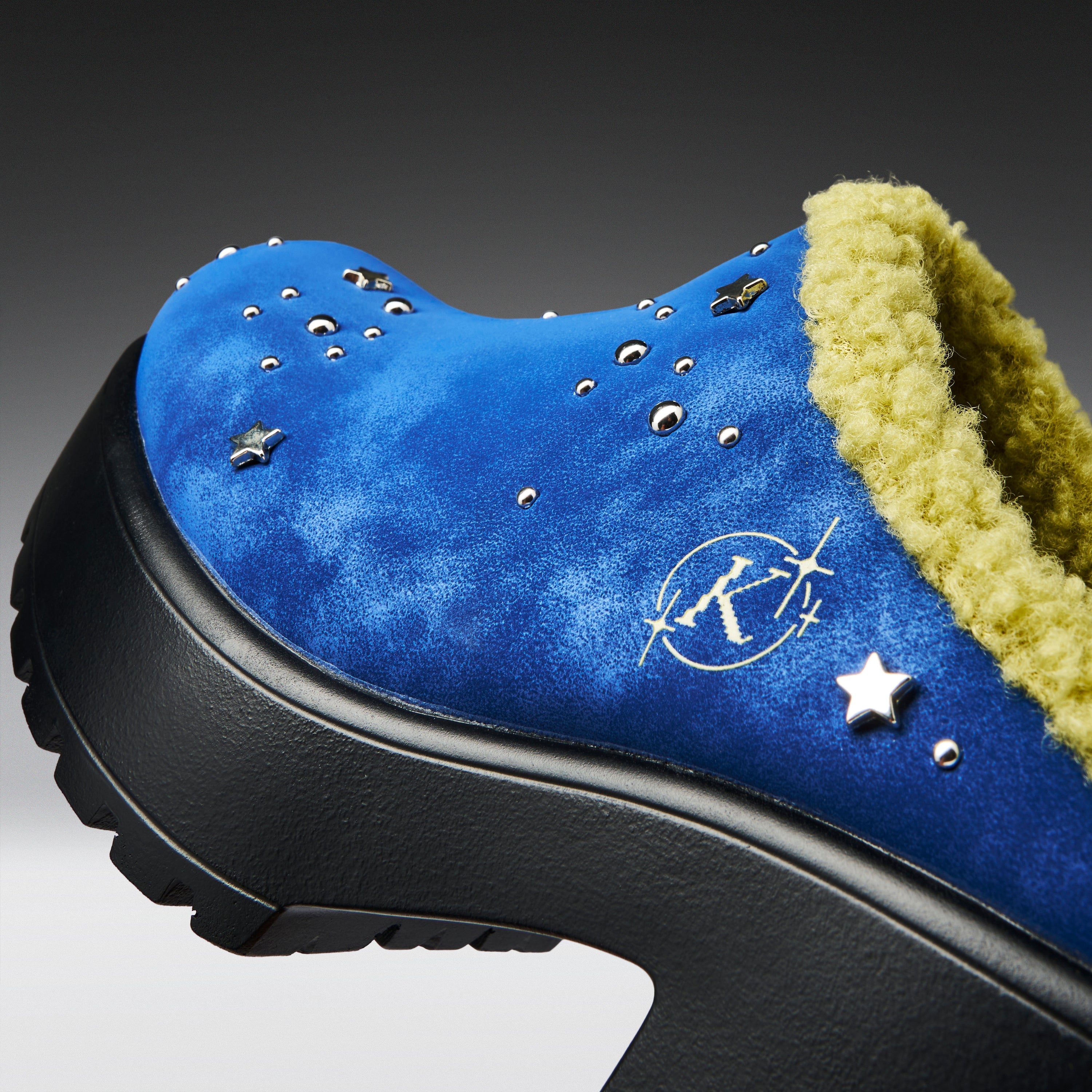 A close-up of a blue suede clog-style shoe with a black platform sole, a yellow-green sherpa lining, and silver star and stud embellishments.