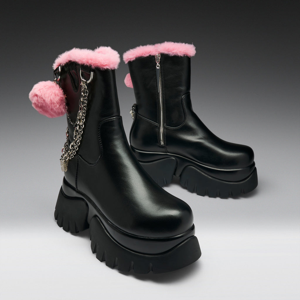 A pair of black leather ankle boots with pink faux-fur trim, chunky black platform soles, and a pink pom-pom charm with a silver chain on the side, set against a dark gray gradient background.