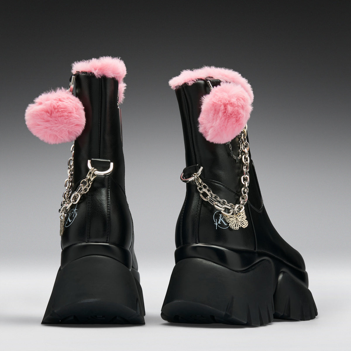 A pair of black ankle boots with fluffy pink faux-fur trim, pink pom-poms, and silver chain details, shown from the back on a gray gradient background.