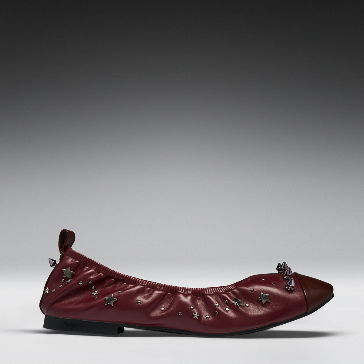 A single burgundy leather ballet flat with star-shaped metallic embellishments and a dark brown pointed toe, shown in profile against a gray gradient studio background.