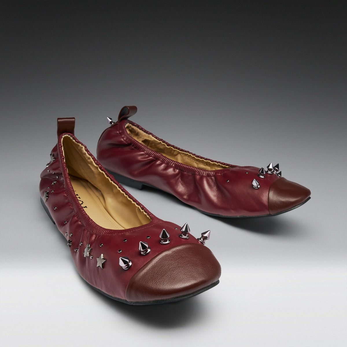 A pair of burgundy leather ballet flats with metallic silver spikes and star-shaped studs, displayed on a gray gradient studio background.
