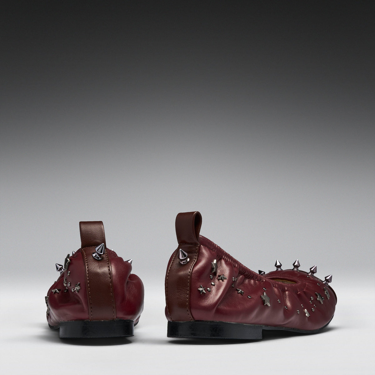 A pair of burgundy leather ballet flats decorated with silver metal spikes and butterfly embellishments, shown from the back against a dark-to-light gray gradient background.