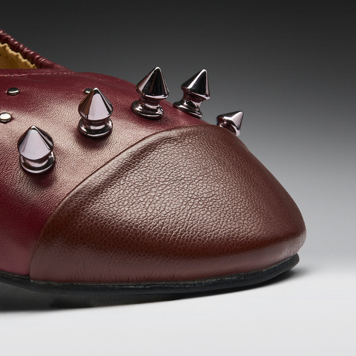 A close-up of a burgundy leather shoe featuring metallic spiked studs on the toe area, set against a dark gray gradient background.