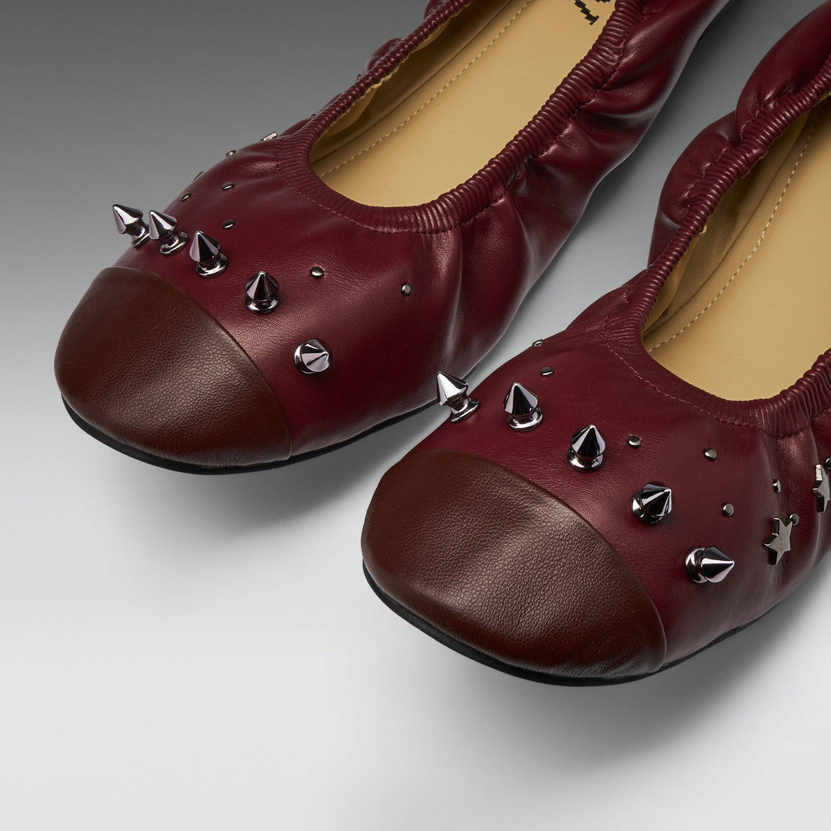 A pair of burgundy leather ballet flats with dark brown toe caps and silver metal spikes, shown on a neutral gray background.