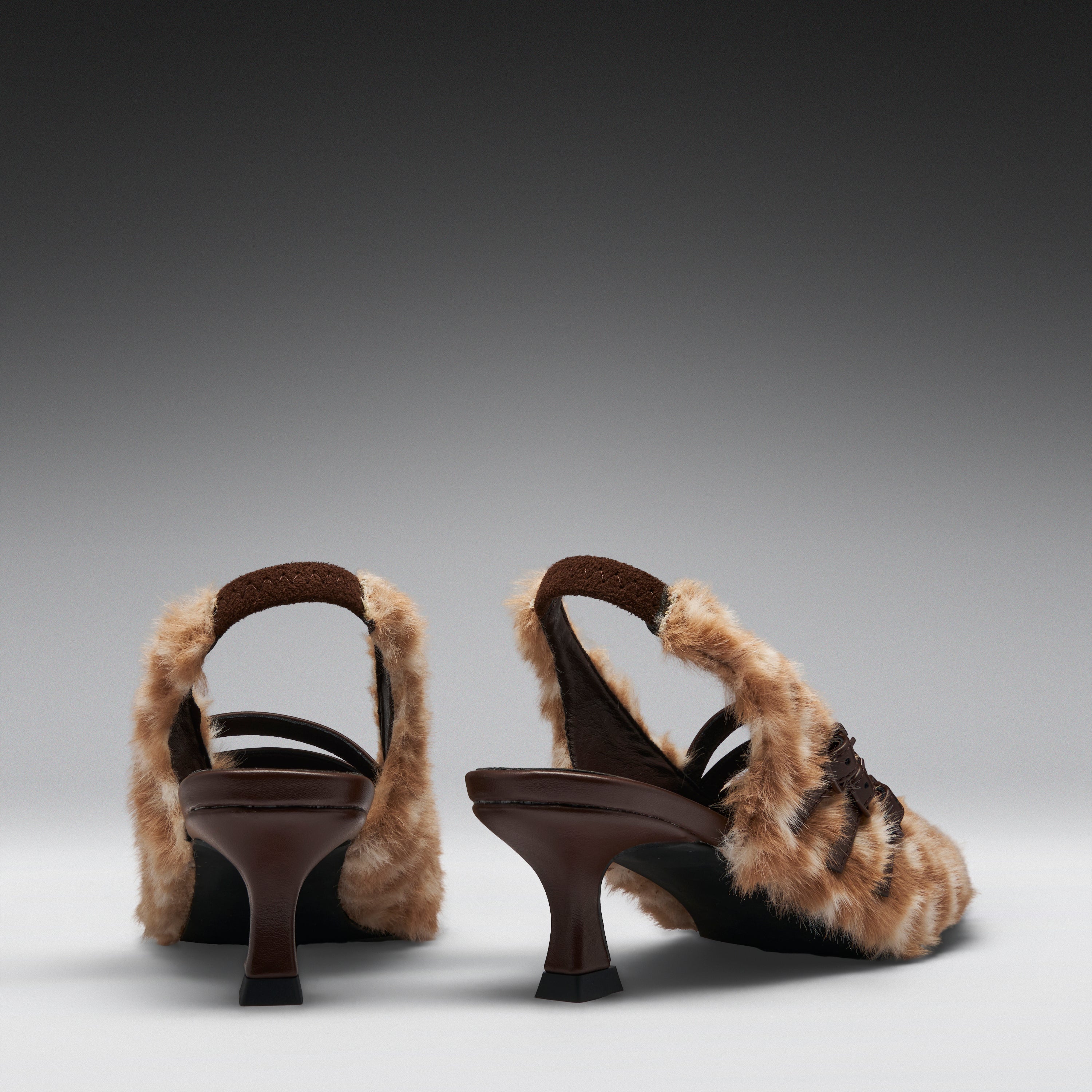 A pair of women's slingback high-heeled sandals featuring brown straps and thick, fluffy beige faux-fur uppers, shown from a rear three-quarter angle on a dark-to-light gray gradient studio background.