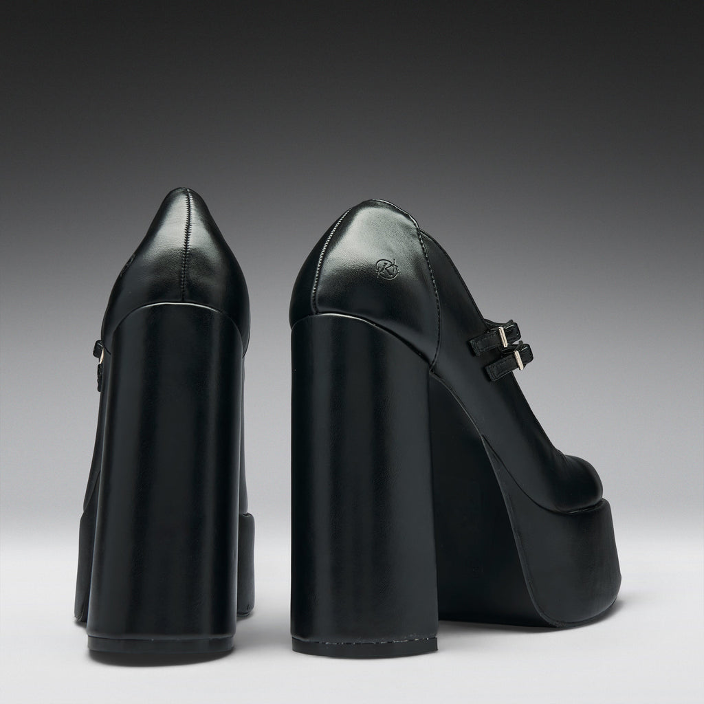 A pair of black leather high-heeled pumps with chunky block heels and platform soles, shown from the back and side against a dark gray gradient background.