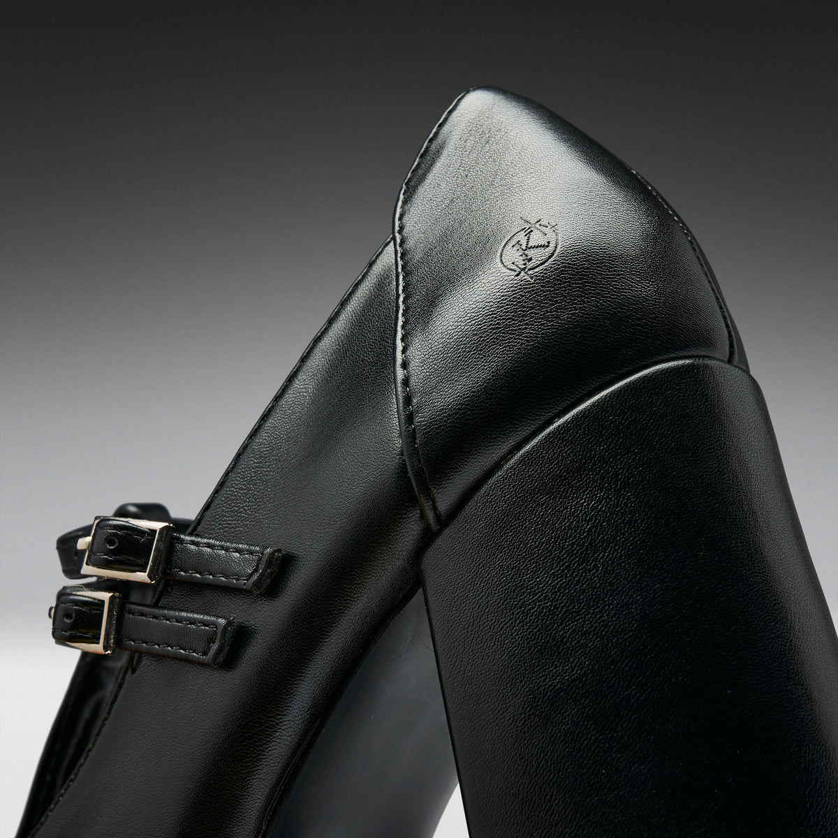 Close-up of a black leather boot with double buckle straps and a small embossed logo on the heel, photographed against a dark gray gradient background.