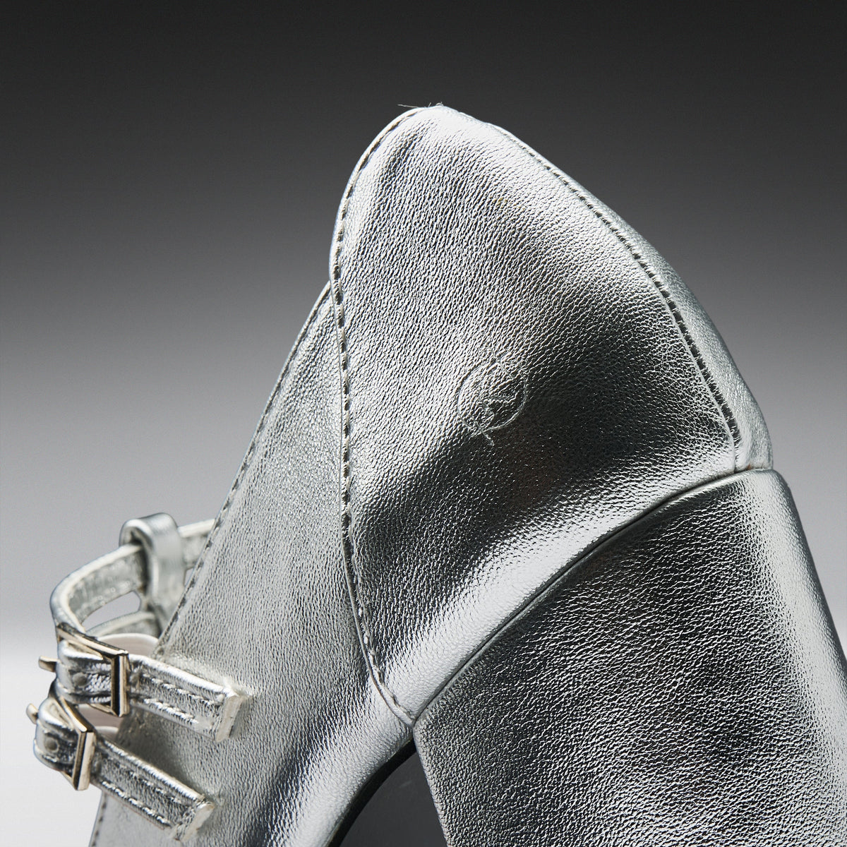 Close-up of a metallic silver leather shoe with a pointed toe and double strap detailing on a dark gray gradient background.