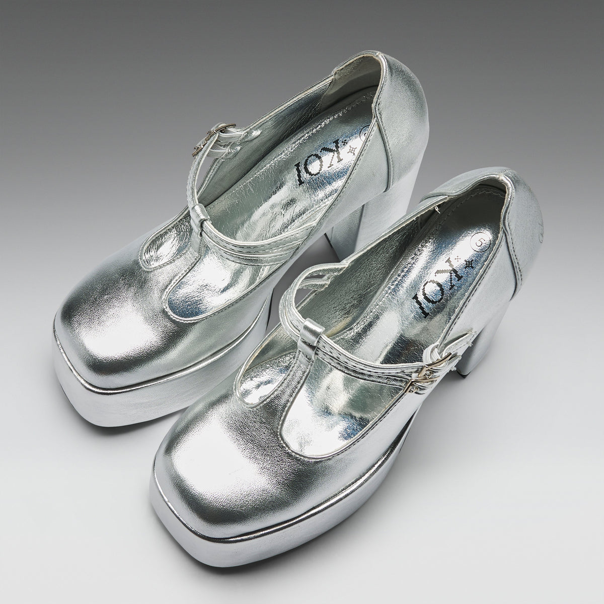 A pair of metallic silver Mary Jane platform heels with block heels and buckle straps, displayed on a neutral gray gradient background. The brand name 'KOT' is visible on the insoles.