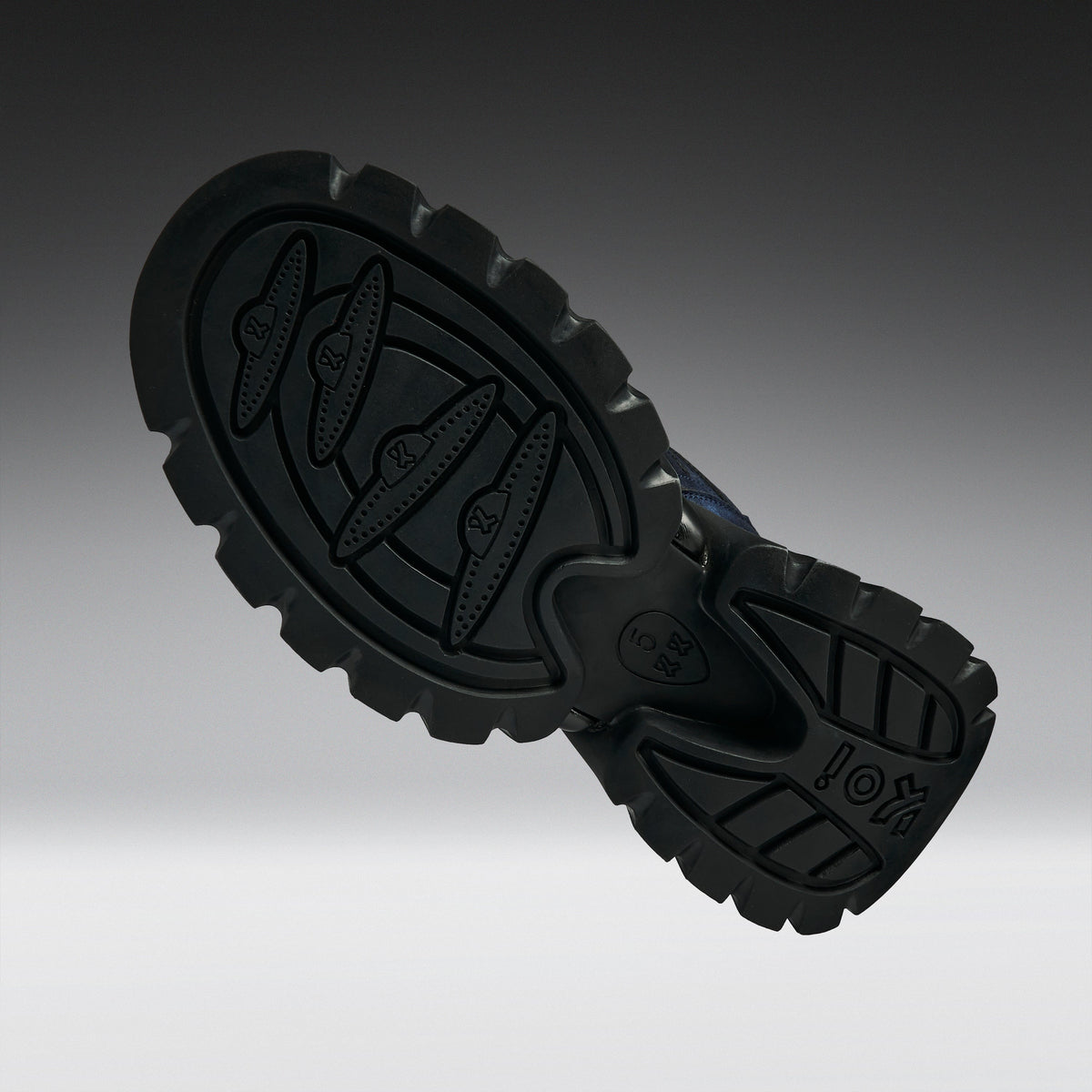 A close-up of a black rubber shoe sole with deep tread patterns and embossed logo details, shown against a dark-to-light gray gradient background.