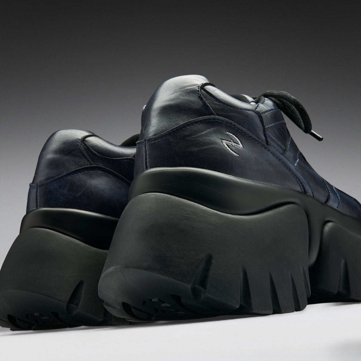 A pair of chunky low-top sneakers in dark navy blue leather with thick black platform soles, shown from a low angle against a dark gray gradient background.