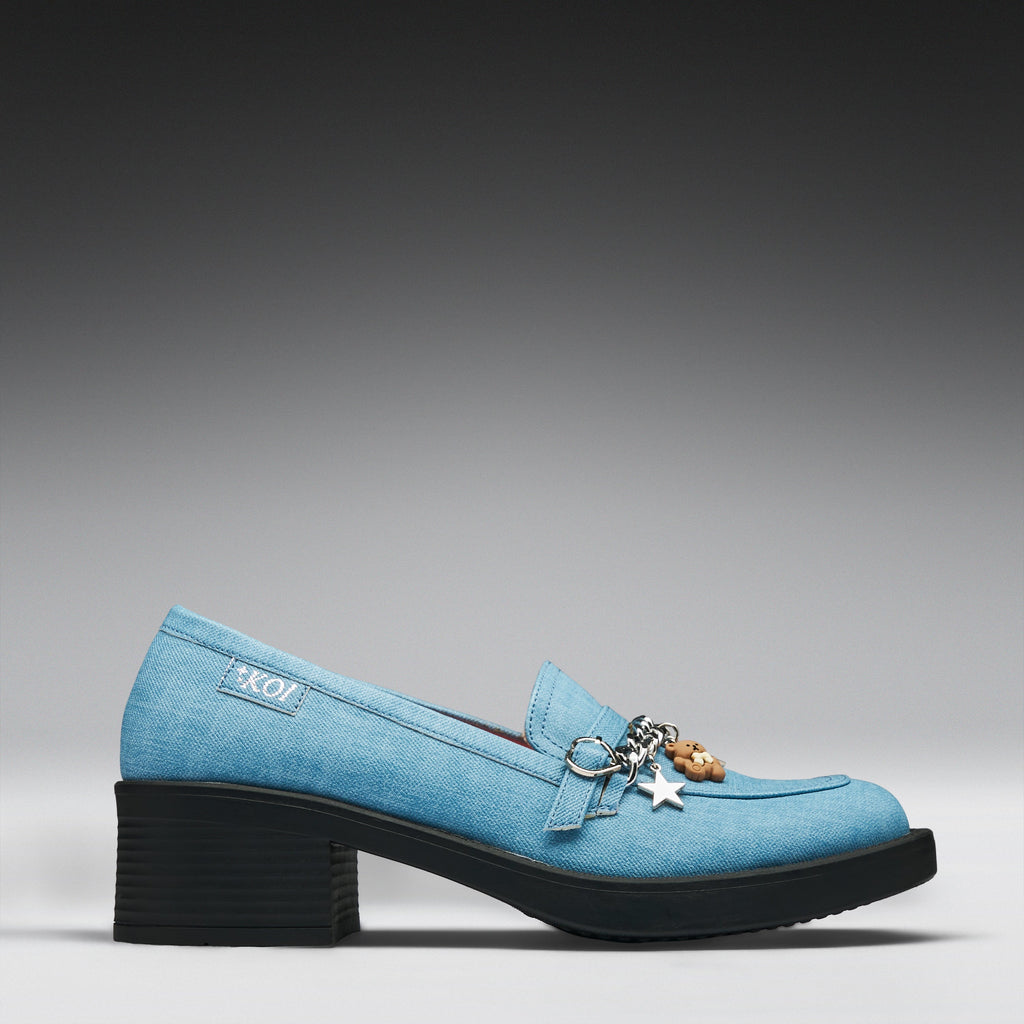 A light blue denim-textured loafer-style shoe with a chunky black block heel, decorative metal rings, and hanging charms, shown in side profile on a gray gradient background.