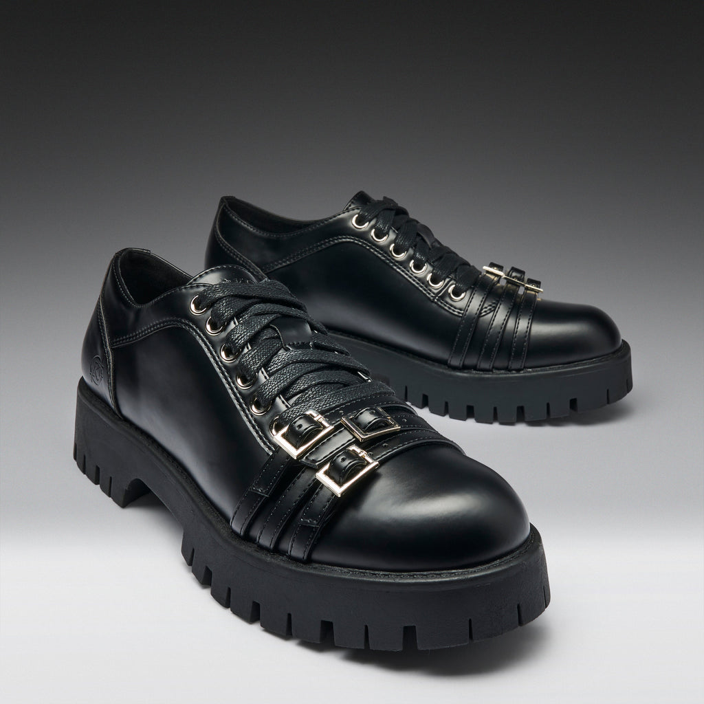 A pair of black leather lace-up shoes with chunky lug soles and decorative gold-tone buckle straps on the sides, photographed on a dark-to-light gray gradient studio background.