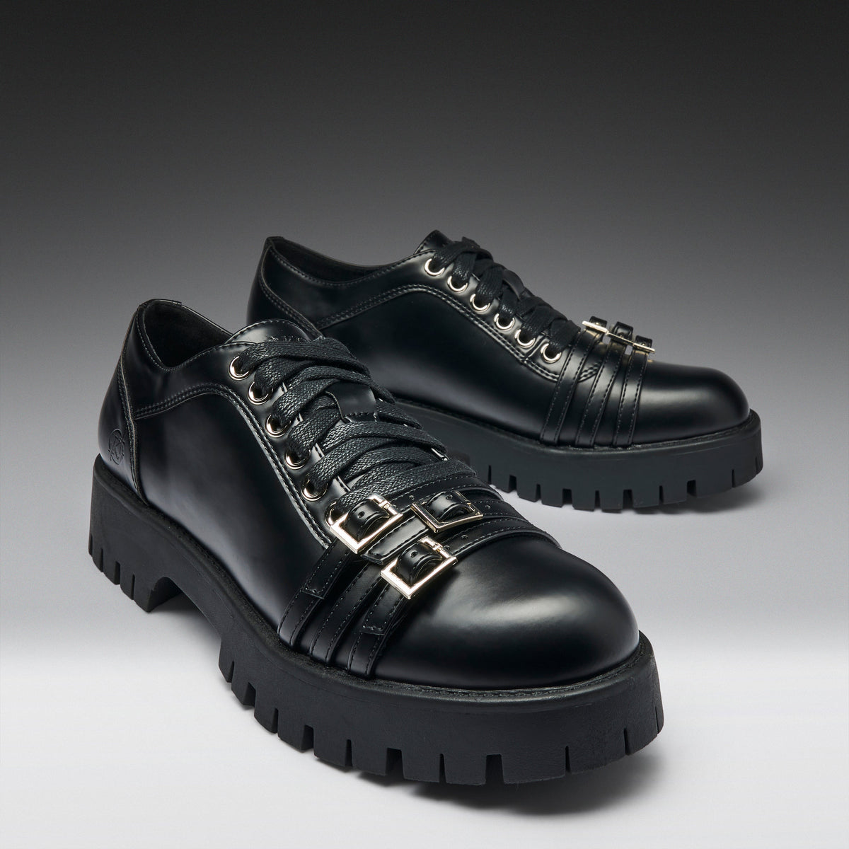 A pair of black leather lace-up shoes with chunky lug soles and decorative gold-tone buckle straps on the sides, photographed on a dark-to-light gray gradient studio background.