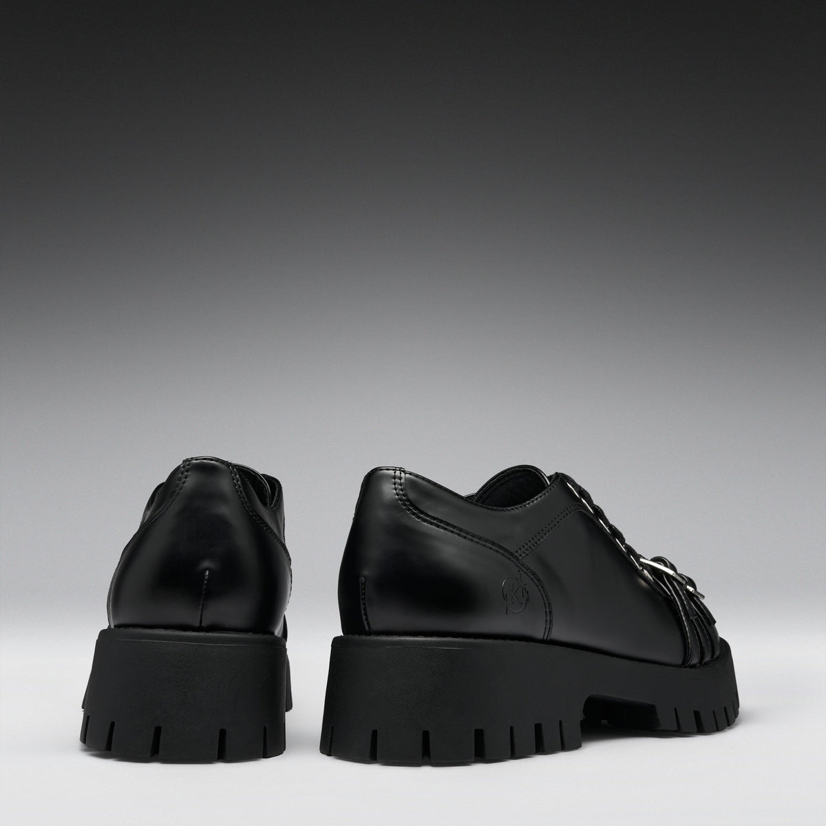 A pair of black leather lace-up shoes with chunky lug soles, shown from the back and side against a dark gray gradient background.