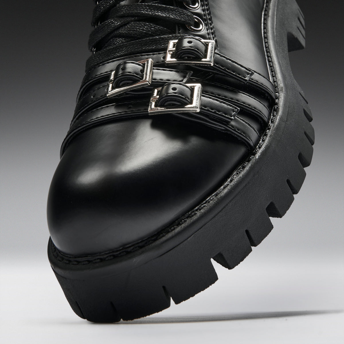 Close-up of a black leather lace-up boot with two silver buckle straps and a chunky lug sole, photographed on a gray studio background.