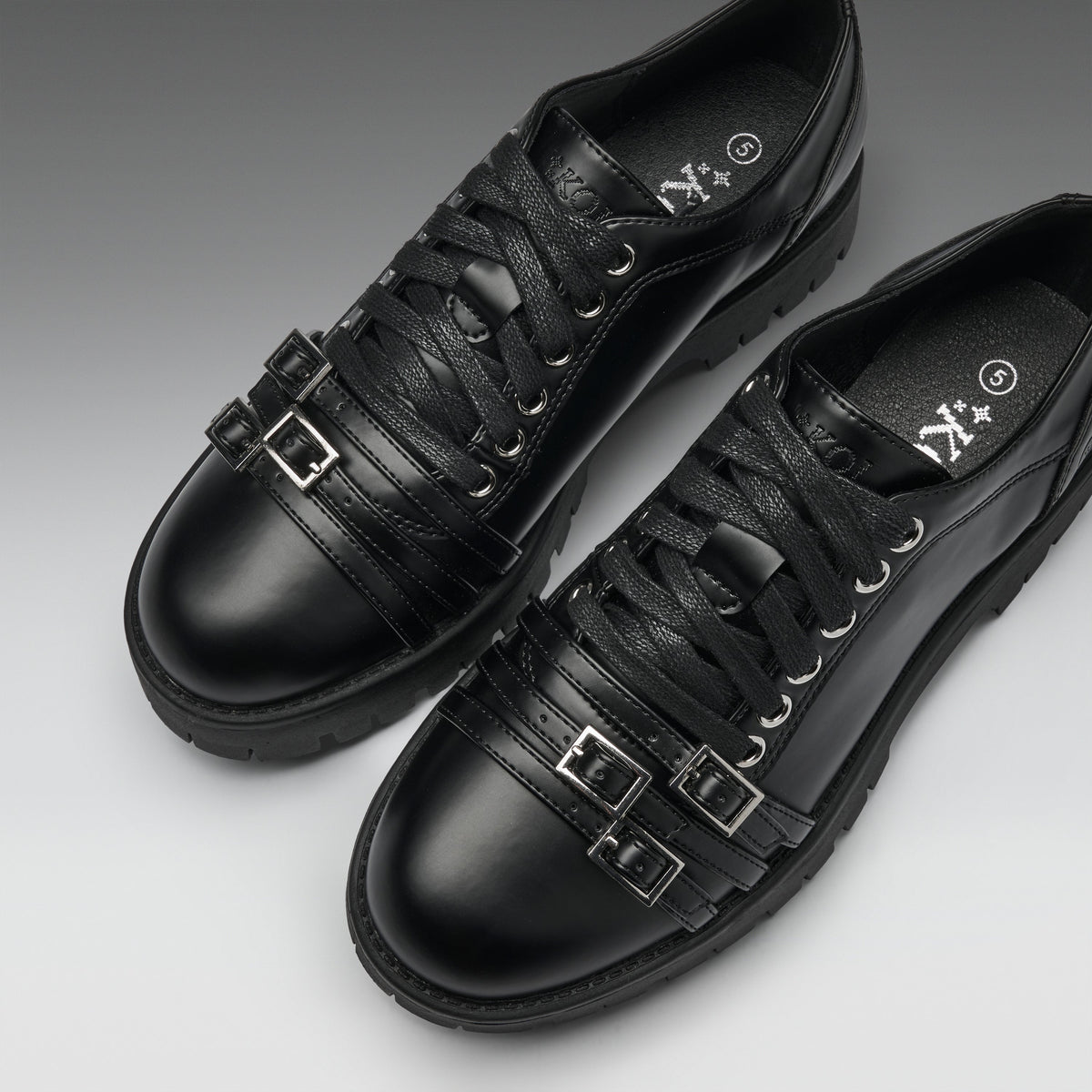 A pair of black leather lace-up shoes with decorative buckle straps on the sides, shown on a clean gray gradient background.