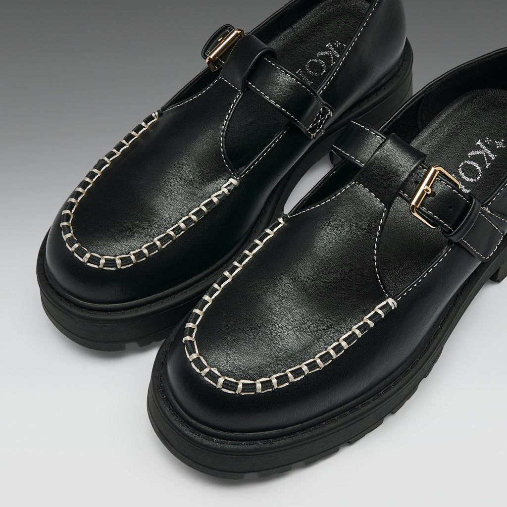 A pair of black leather Mary Jane-style shoes with chain-stitched detailing around the toe, a strap across the instep with a gold-tone buckle, and chunky black soles, photographed on a clean light gray background.
