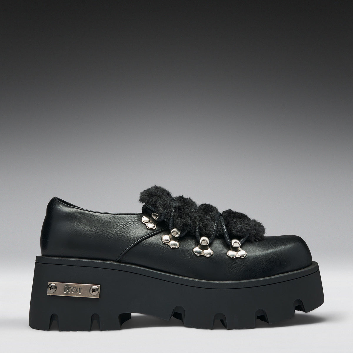 A black leather platform shoe with fluffy black trim, silver-tone metal eyelets, and a chunky lug sole. The brand name 'KOR' is visible on a metal badge on the side of the sole.