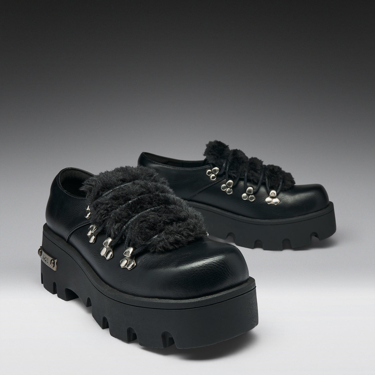 A pair of black leather platform mule shoes with fluffy black faux-fur trim and silver-tone decorative studs, shown on a dark-to-light gray studio background.