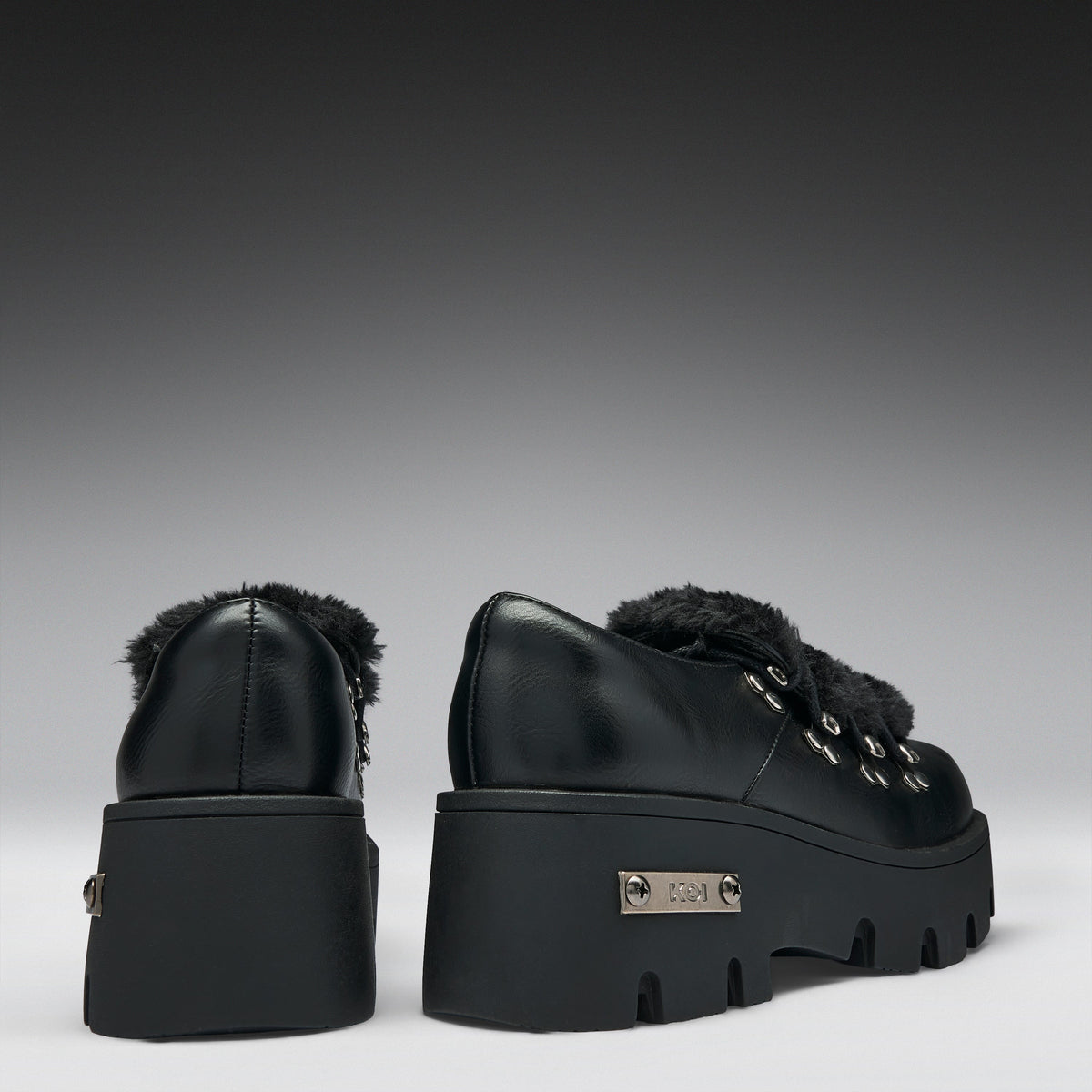 A pair of black leather platform ankle boots with fluffy black faux-fur trim and silver-tone hardware, shown from the back on a dark-to-light gray gradient studio background.
