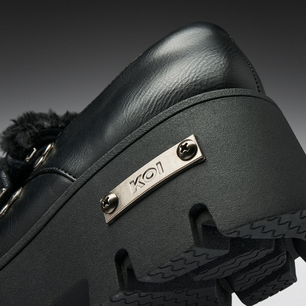 Close-up of a black leather shoe with a chunky black sole, a black furry trim, and a silver metal plate engraved with 'KO1'.