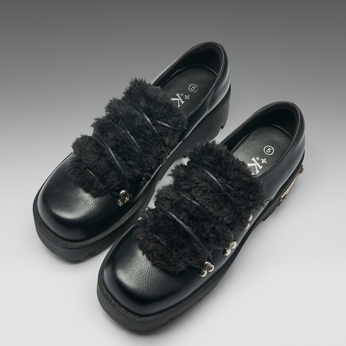A pair of black leather platform loafers with wide, fluffy black faux-fur straps and small silver metal accents, photographed on a neutral gray gradient background.