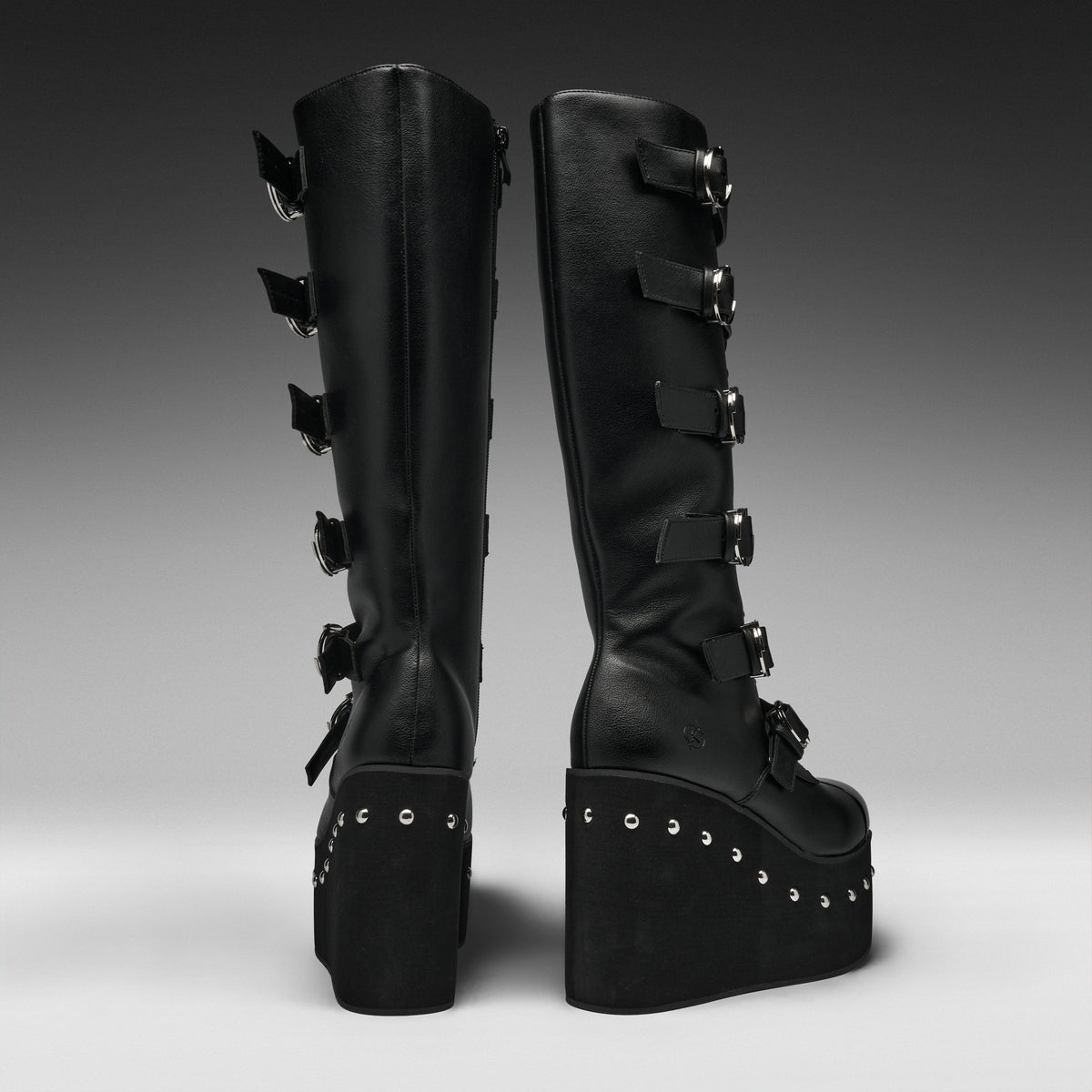A pair of black knee-high platform boots with multiple buckle straps down the side, shown from the back against a dark gray gradient studio background.