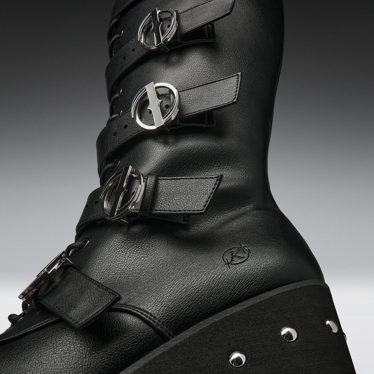 A close-up studio shot of a black leather high-top boot with multiple strap closures and prominent silver-tone buckle hardware, set against a dark gray gradient background.