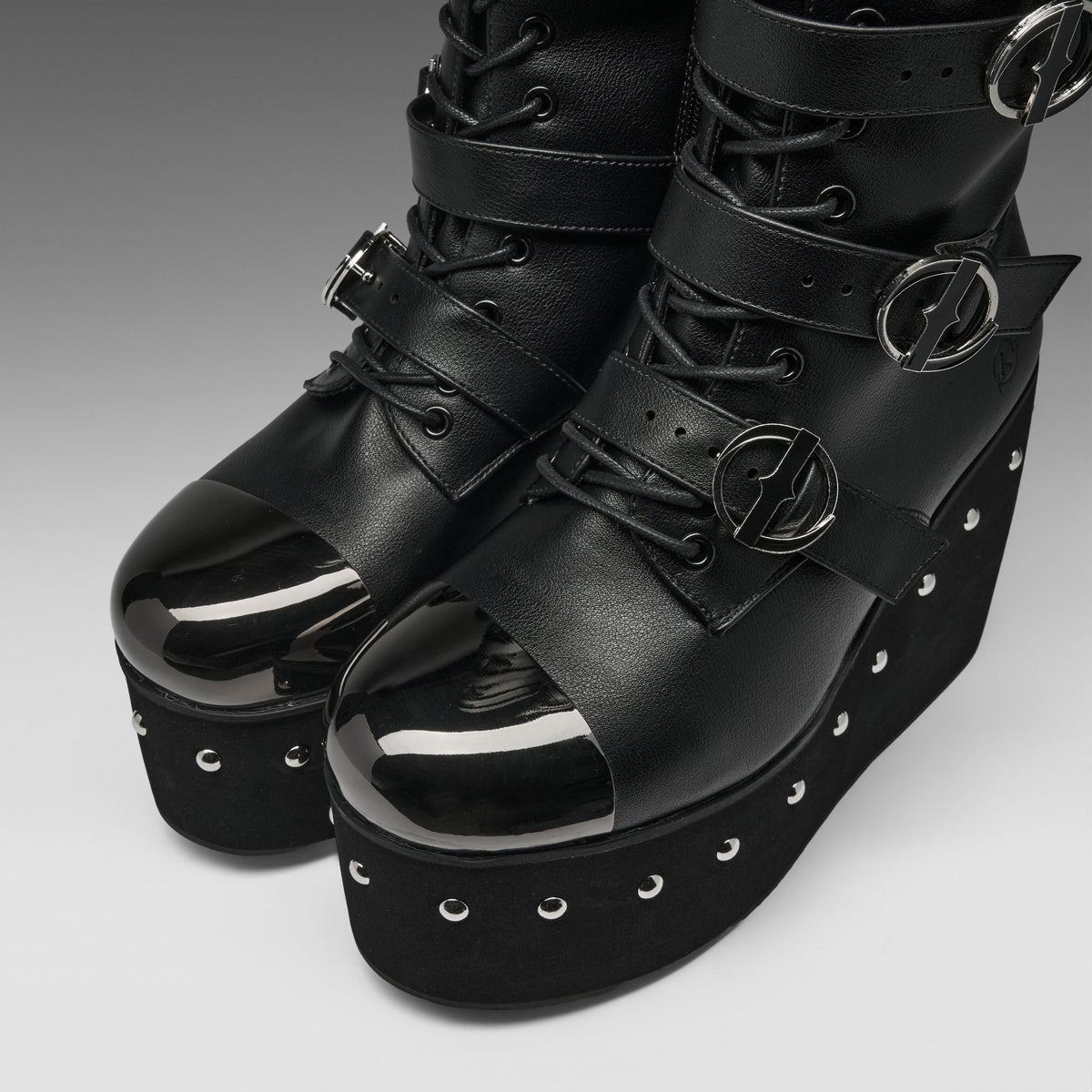 A pair of black platform ankle boots with glossy patent leather toe caps, multiple decorative straps, silver buckle details, and metal eyelets on a gray gradient background.