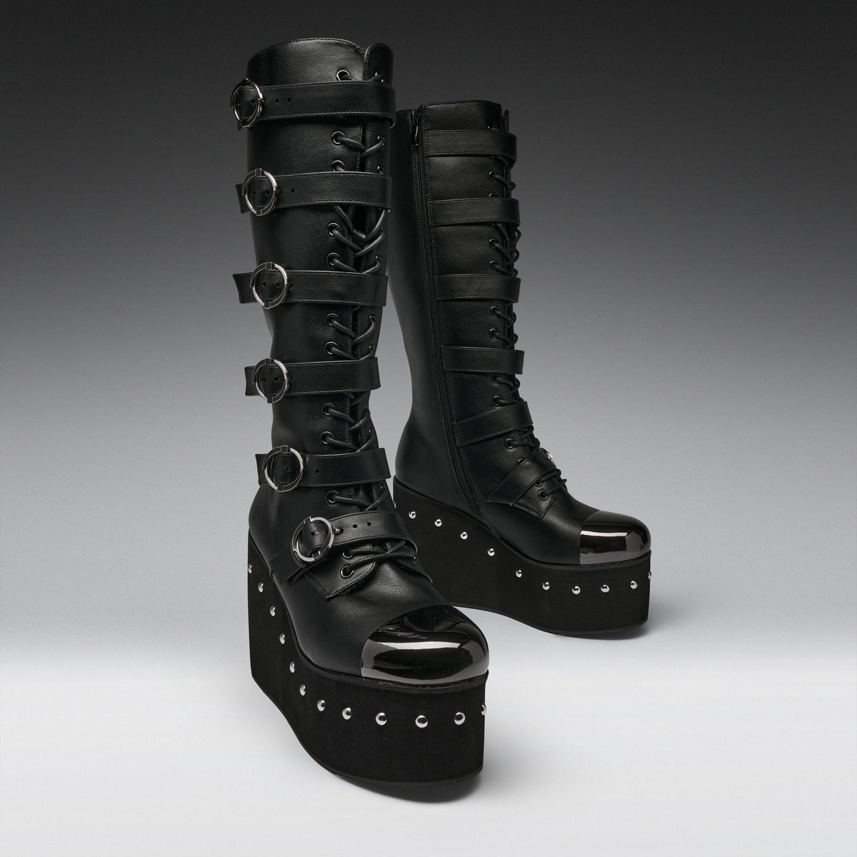 A pair of black platform knee-high boots with multiple buckle straps, lace-up front, and silver studded details, photographed on a dark-to-light gray gradient studio background.