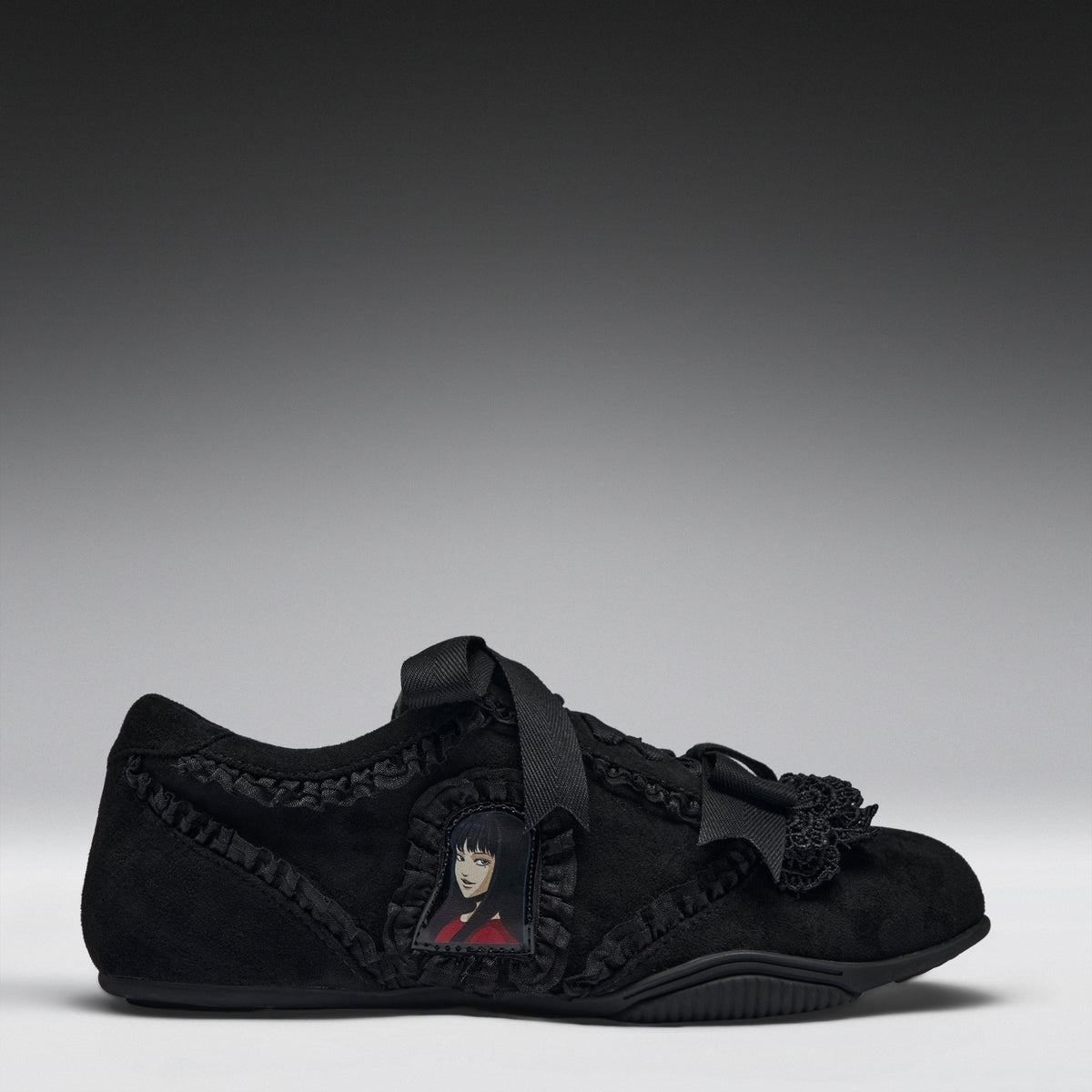 A black suede slip-on shoe featuring a decorative black ribbon strap, ruffled trim, a black tassel accent, and a small anime character patch on the side, set against a dark gray gradient background.