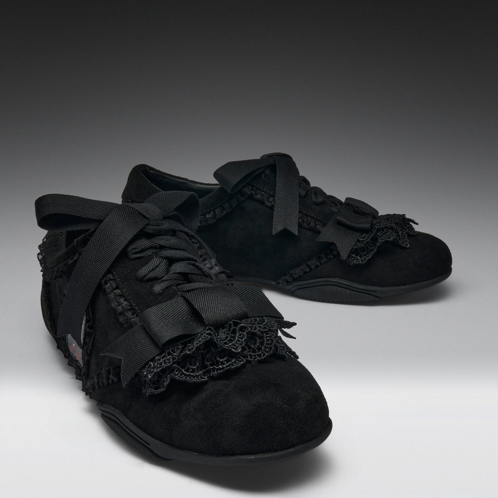 A pair of black lace-up shoes with wide black ribbon bows, black lace trim, and textured black uppers, photographed on a dark-to-light gray studio background.