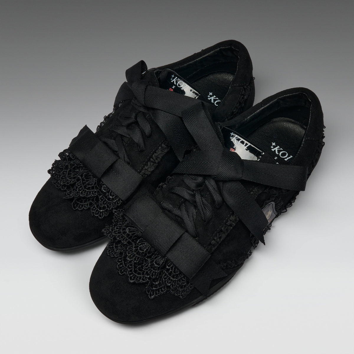 A pair of black lace-up flats with wide satin ribbon straps across the toes and decorative black lace trim on the vamp, photographed on a neutral gray studio background.