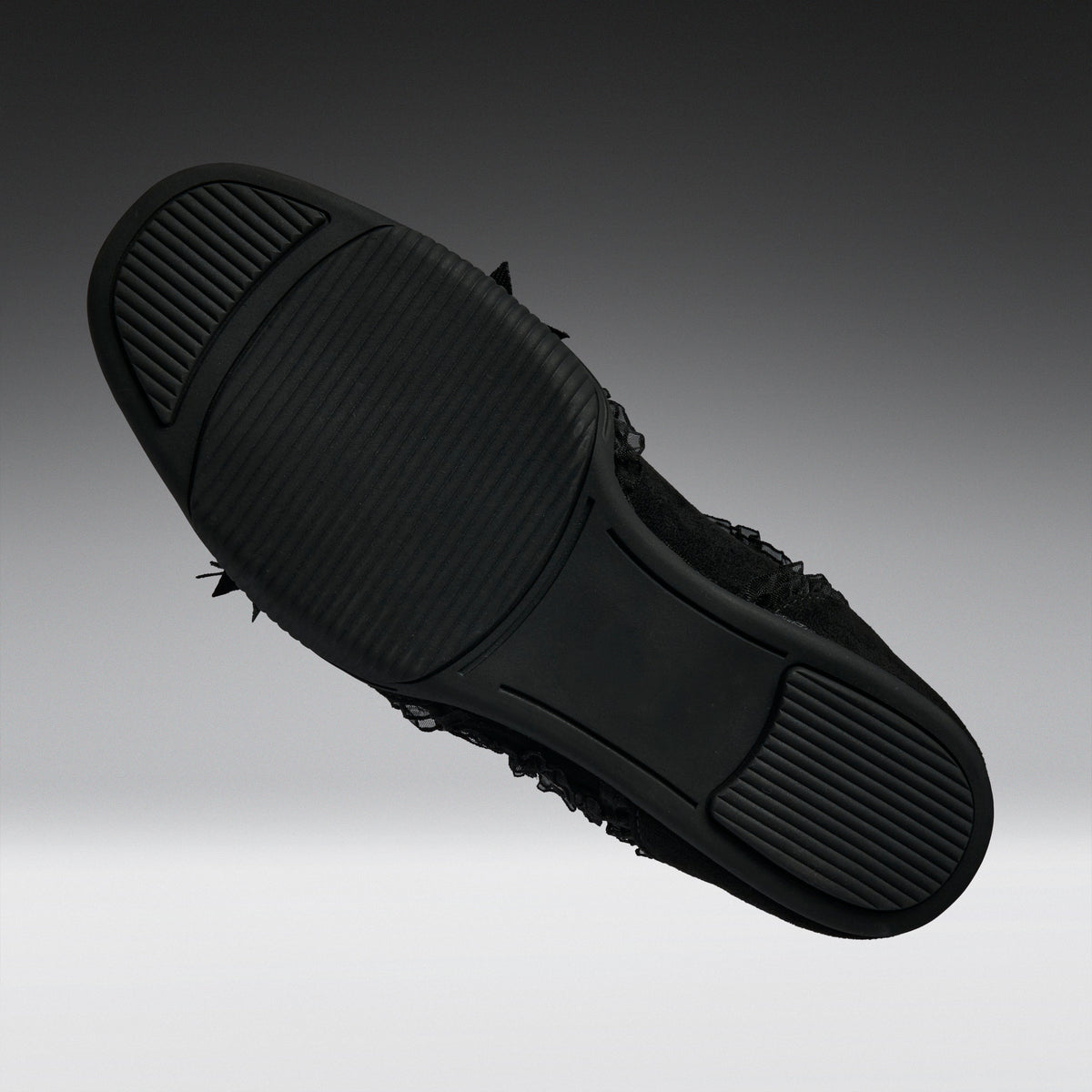 A black slip-on shoe shown from a top-down angle, highlighting its textured rubber sole with small black frayed details along the side, set against a dark gray gradient background.