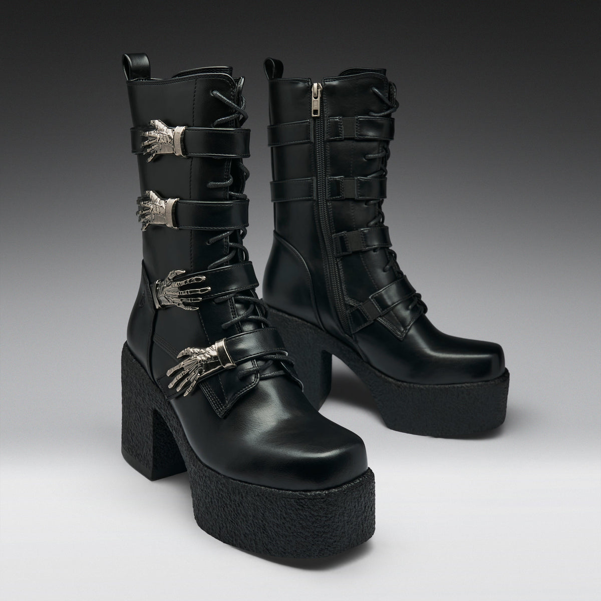 A pair of black leather gothic ankle boots with platform soles, chunky block heels, and silver metallic skeletal hand embellishments, shown against a dark gray studio background.