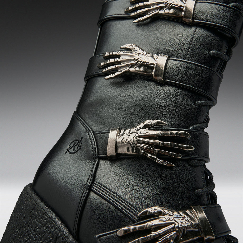 Close-up of a black leather boot with multiple metallic skeleton hand embellishments and silver-toned buckles, shown against a dark gray gradient background.