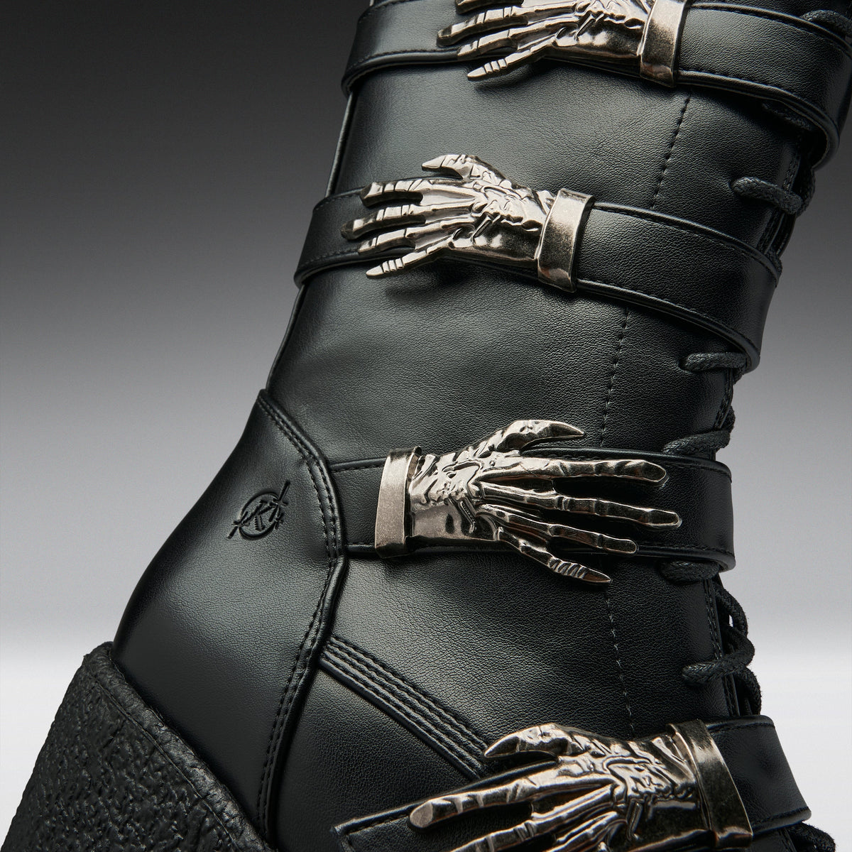 Close-up of a black leather boot with multiple metallic skeleton hand embellishments and silver-toned buckles, shown against a dark gray gradient background.