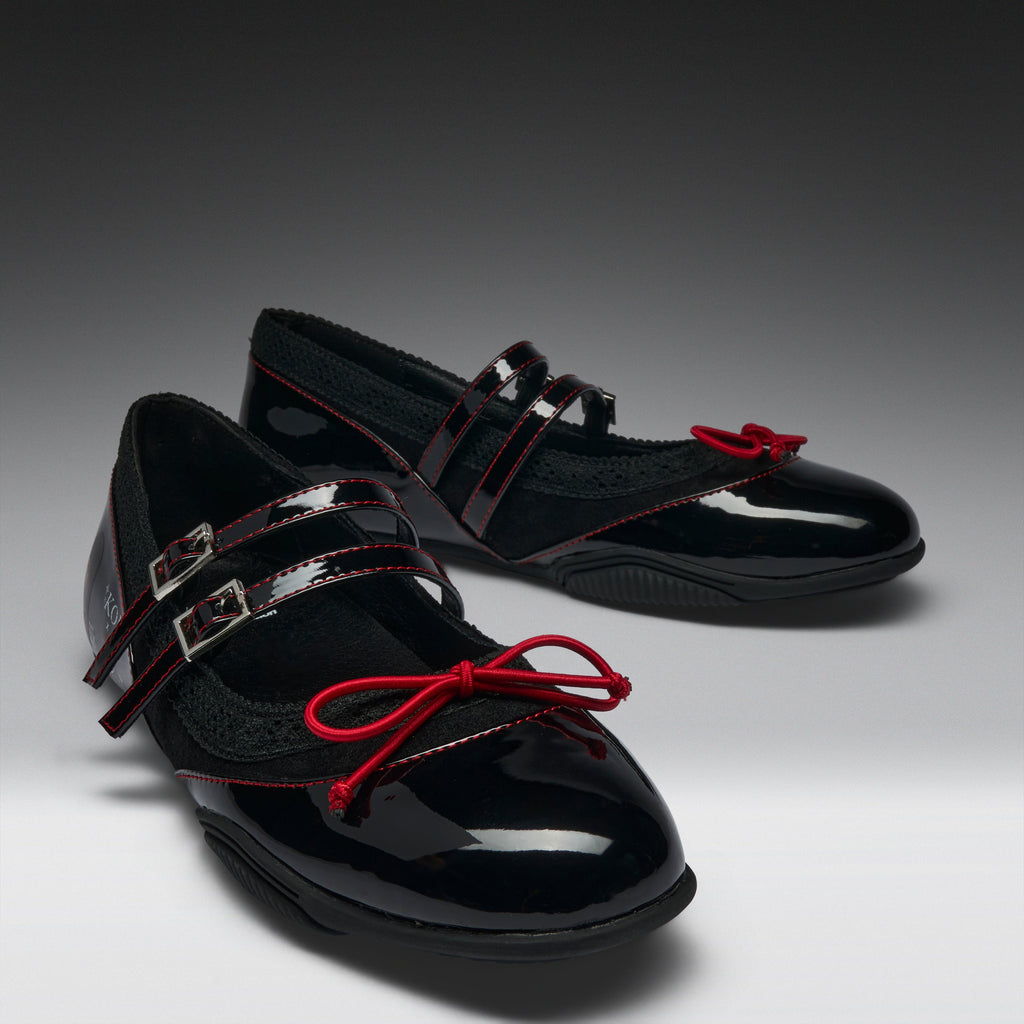 A pair of black patent leather Mary Jane flats with red ribbon bows and red contrast stitching, displayed on a dark gray to white gradient studio background.