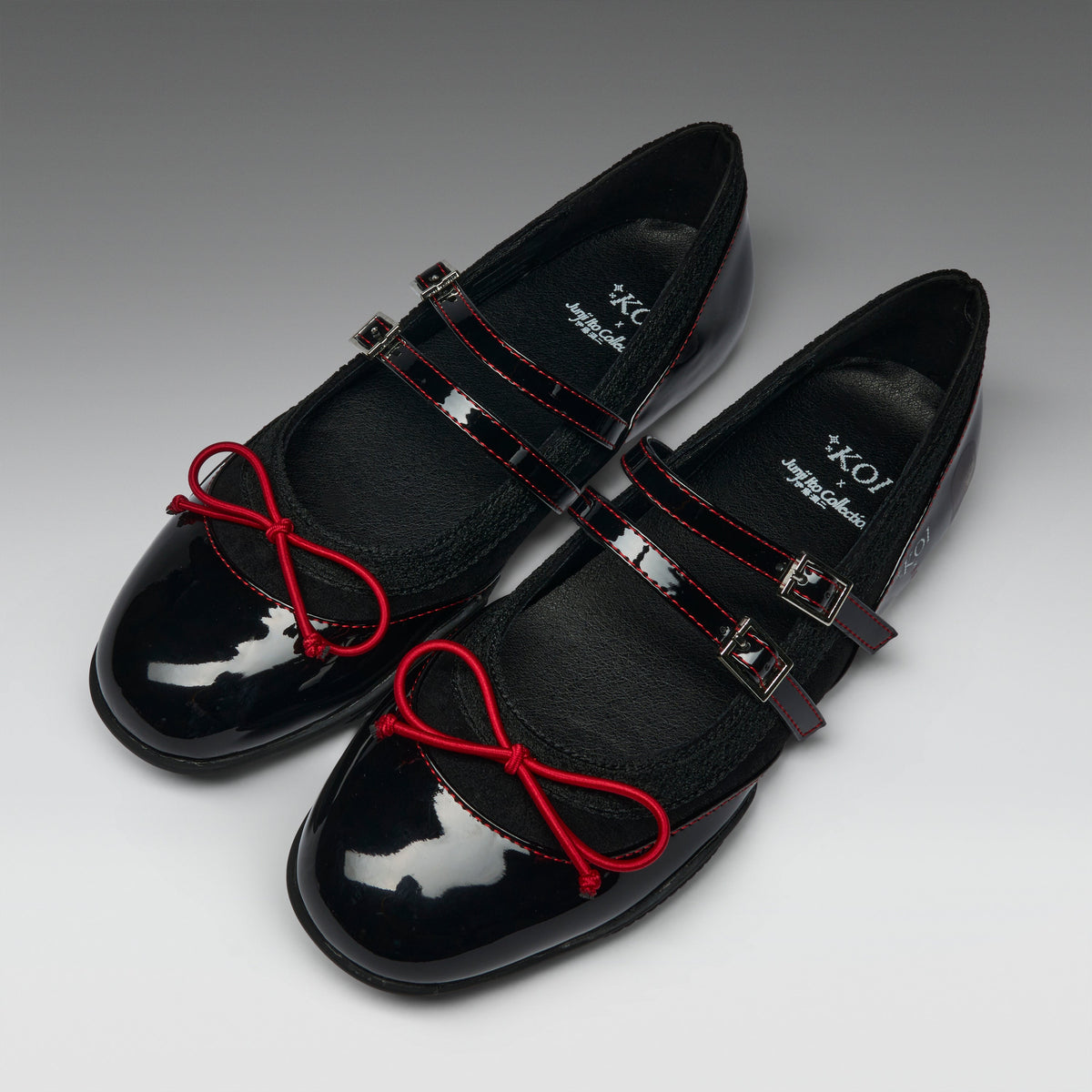A pair of black Mary Jane style flats with glossy patent leather toes, red contrast stitching, red bow details on the straps, and silver buckle closures, photographed on a gray gradient background.