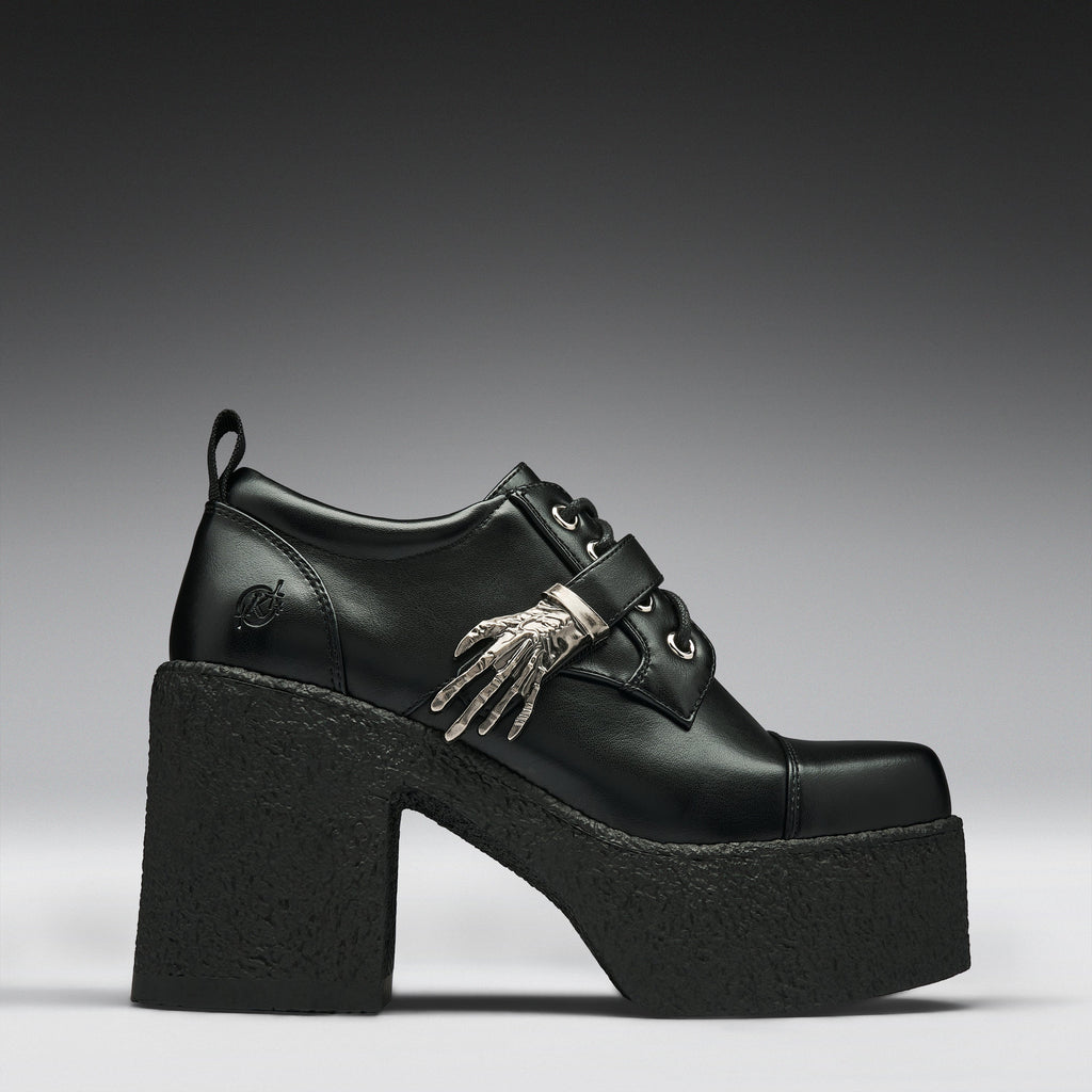 A black leather platform clog shoe with a chunky black sole and heel, featuring a decorative silver skeleton hand ornament on the side, set against a dark gray gradient background.