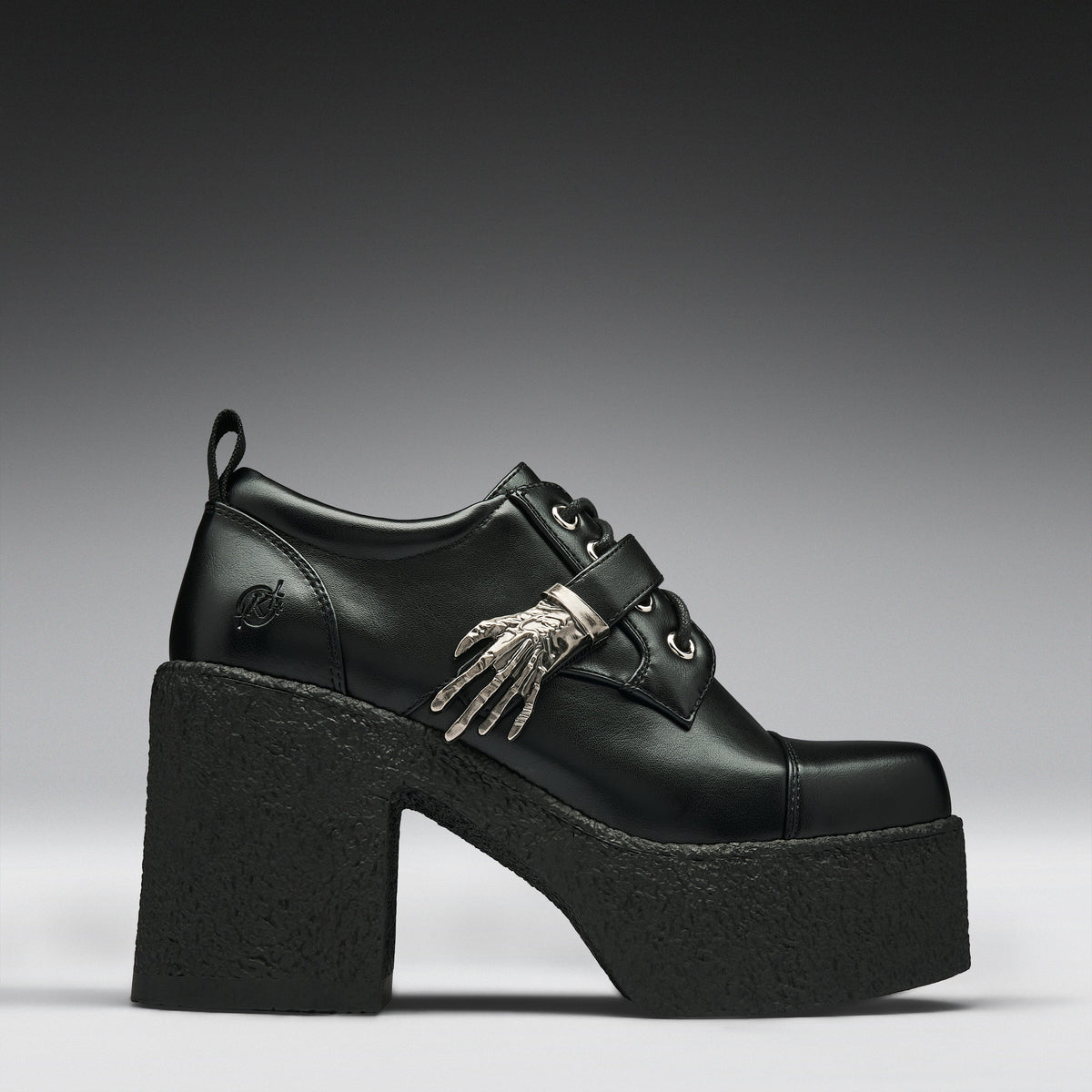 A black leather platform clog shoe with a chunky black sole and heel, featuring a decorative silver skeleton hand ornament on the side, set against a dark gray gradient background.