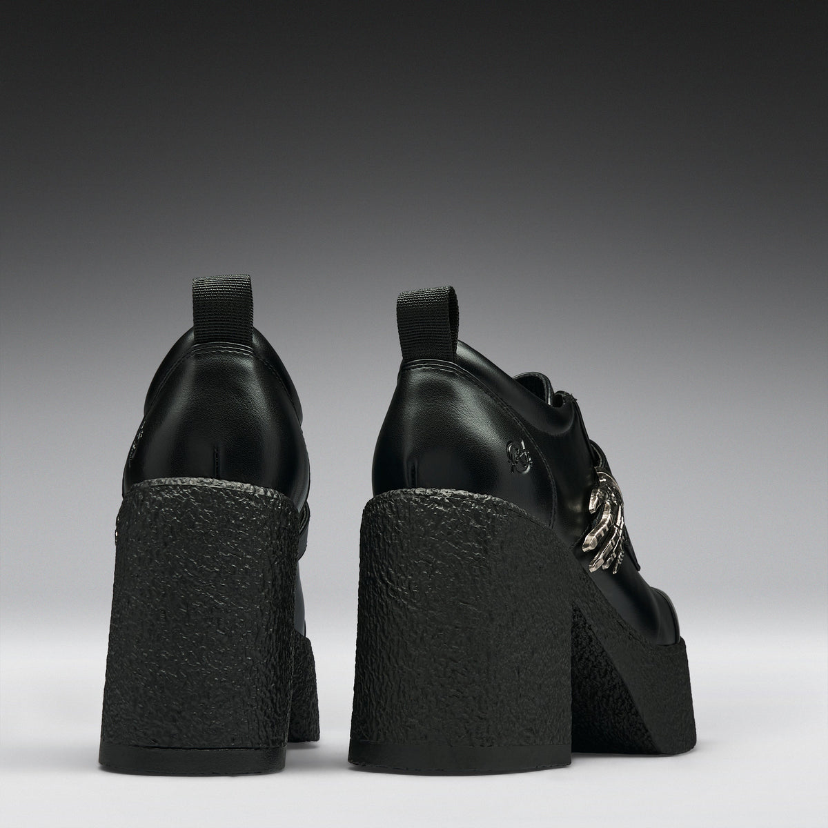 Black leather platform ankle boots with chunky textured block heels and silver chain details, shown from the back on a dark gray gradient background.