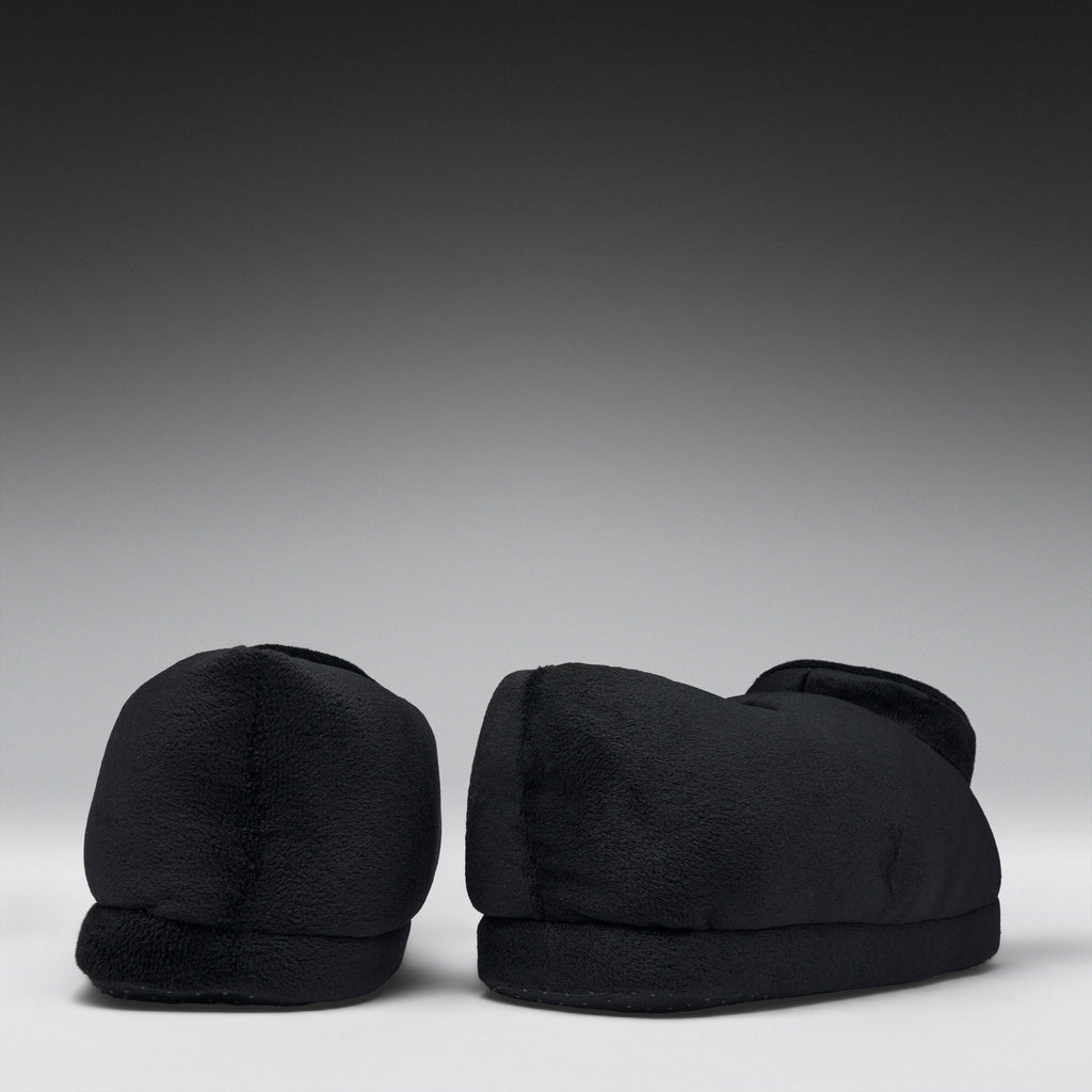A pair of black plush slippers shown from the back against a dark gray gradient studio background.