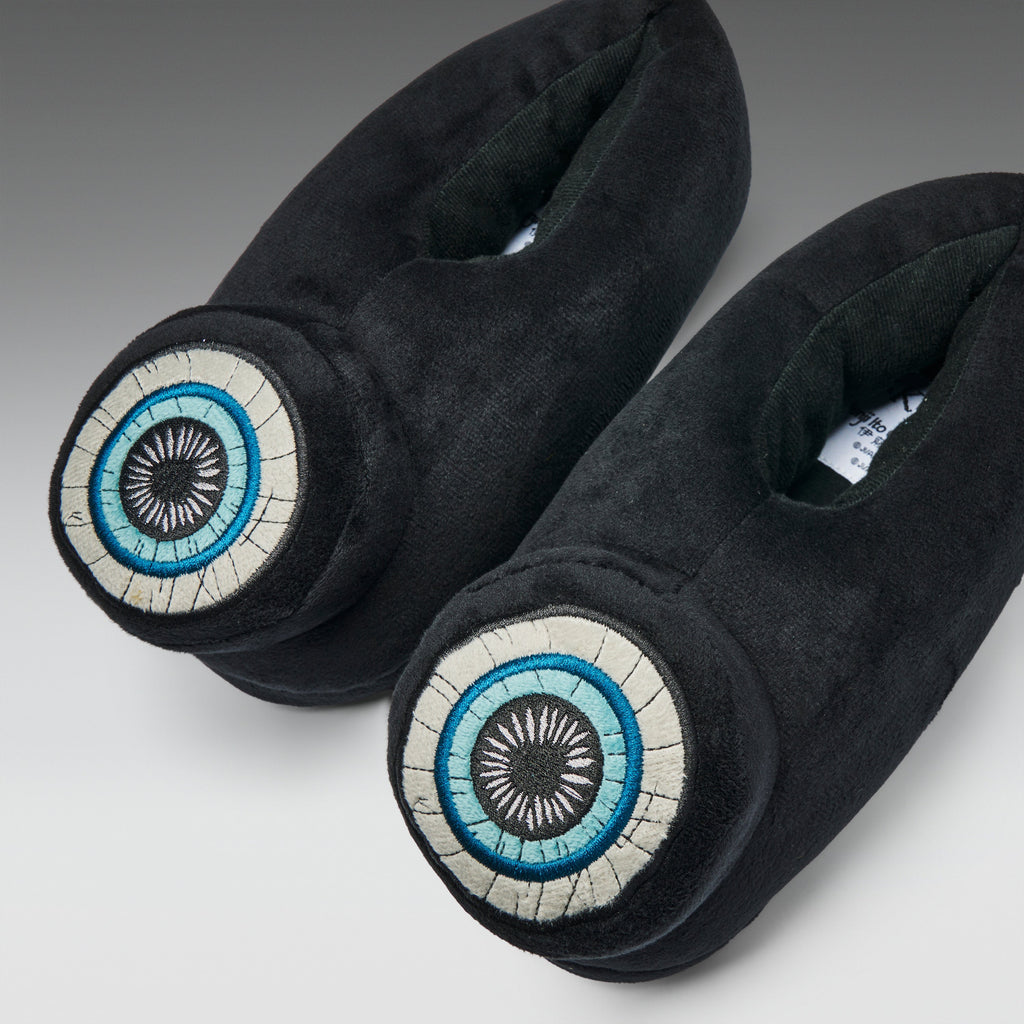 A pair of black plush slippers with circular embroidered eye motifs on the toes, featuring blue, white, and black details, set against a neutral gray background.